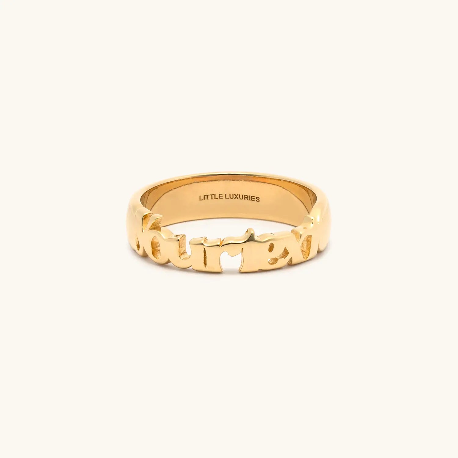 A gold ring with the word 'yourtext' cut out, shown on a light cream background. The inside of the band is engraved with 'LITTLE LUXURIES'.