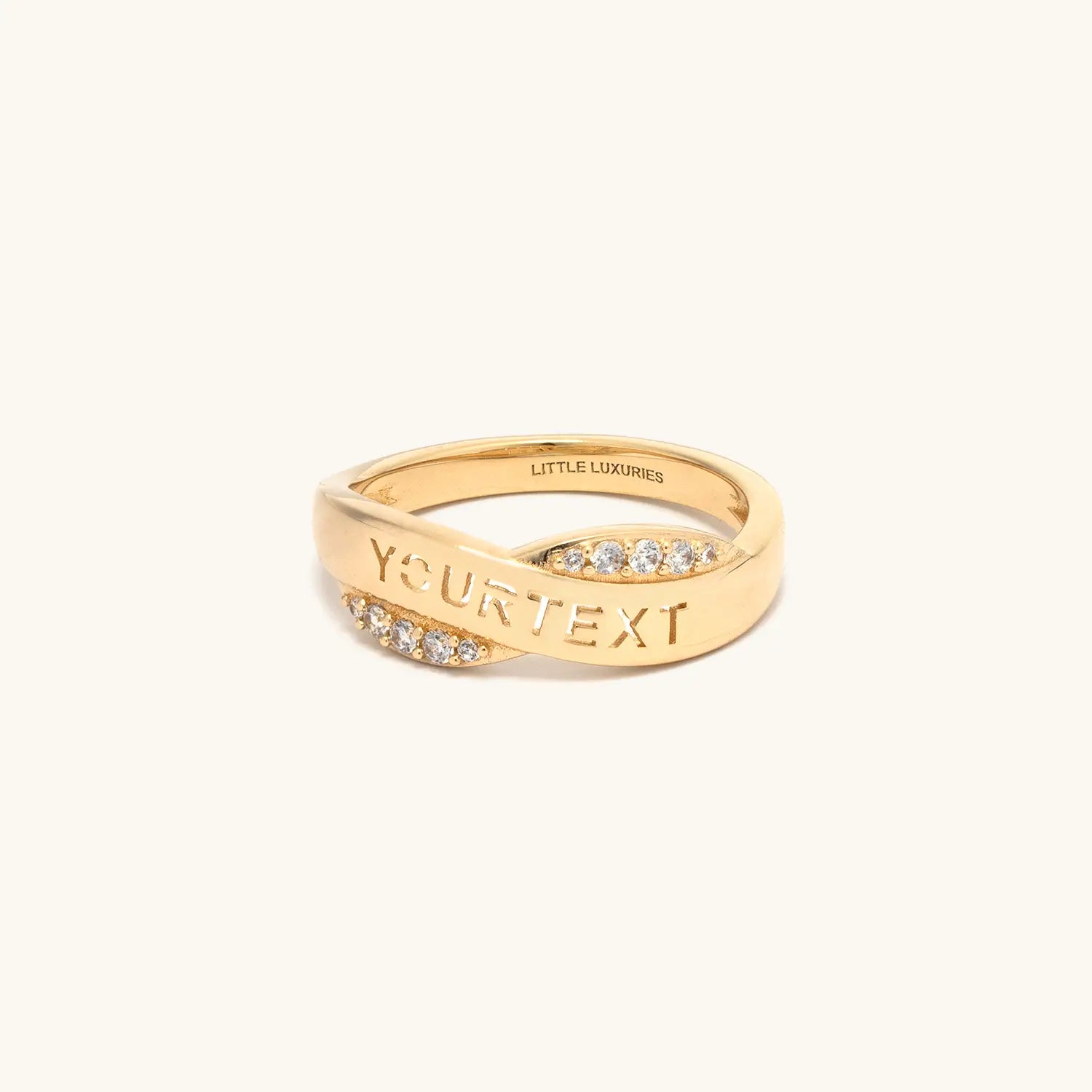 A gold ring with a twisted design, featuring rows of small clear stones on the front and the text 'YOURTEXT' engraved on the band. The brand name 'LITTLE LUXURIES' is visible inside the band.