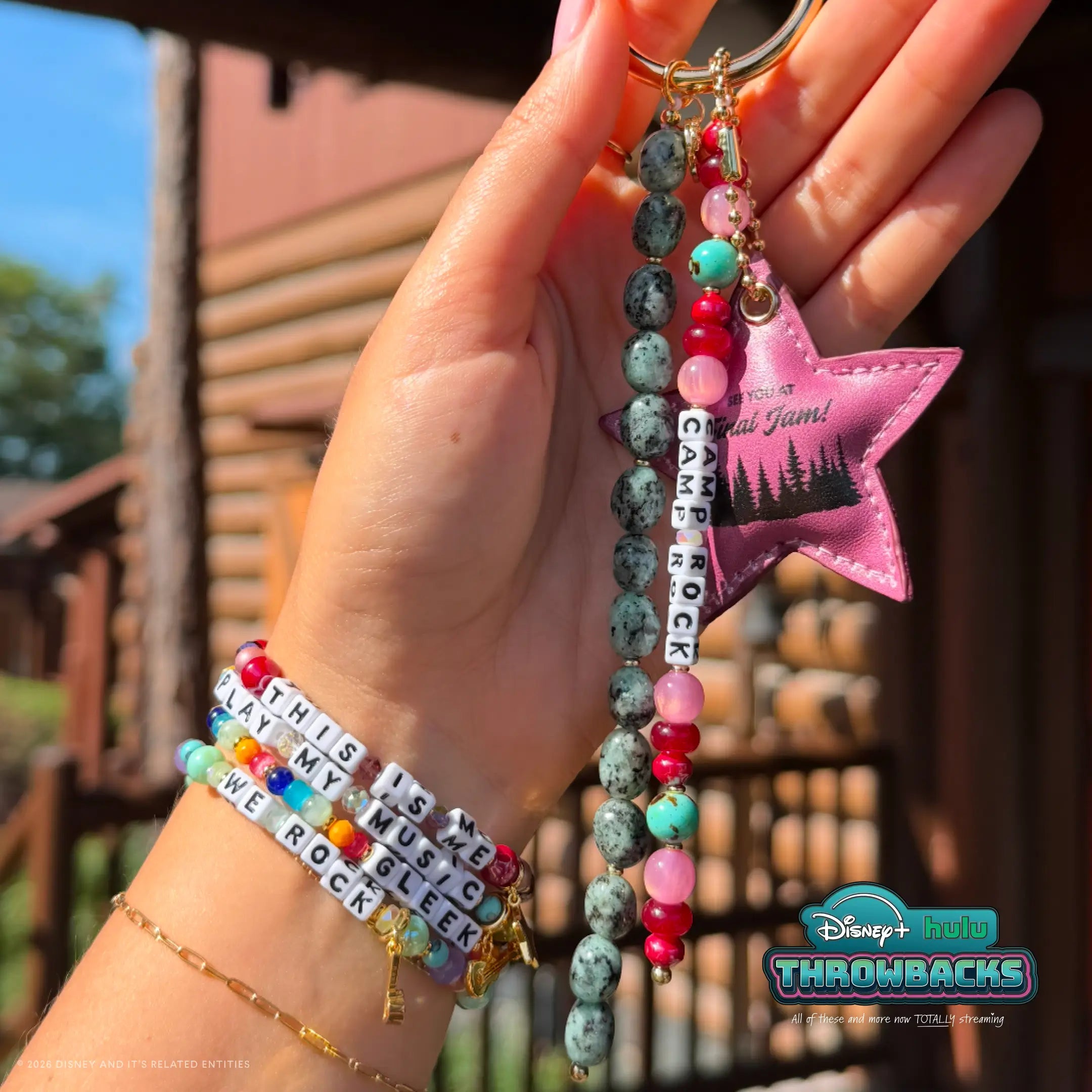 Camp Rock Disney+ Hulu Throwbacks bag charm held in a hand outdoors at a camp lodge, styled with the Throwbacks bracelet stack