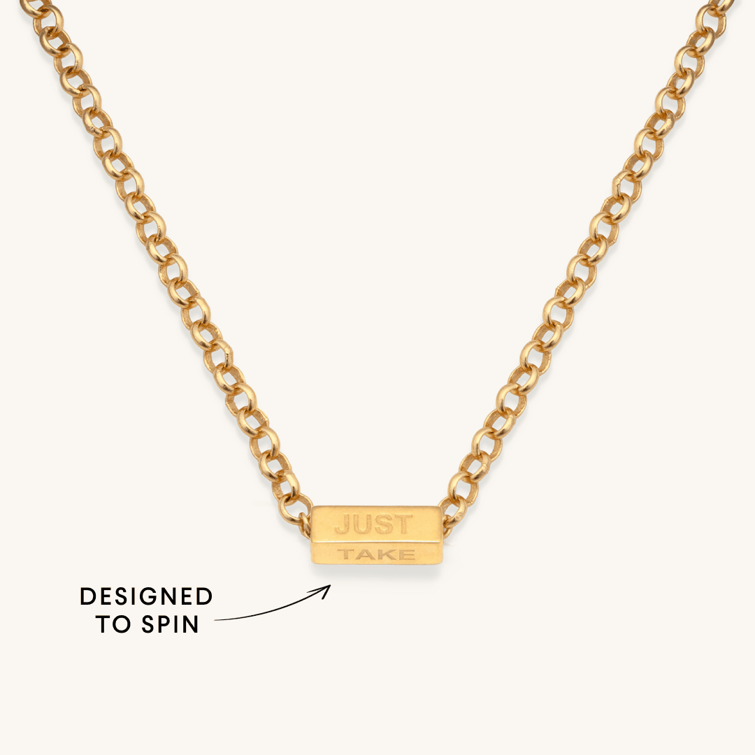Gold curb chain necklace with a spinning "JUST / TAKE" bar charm, designed to spin, on a light background.