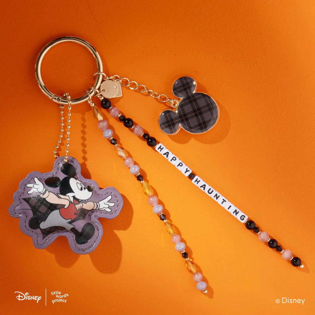 Happy Haunting Disney Mickey & Friends Bag Charm with a faux leather vampire Mickey Mouse figure, plaid Mickey Mouse silhouette charm, and black, pink, and amber beaded Happy Haunting word strand on a gold key ring, flat-lay on orange background