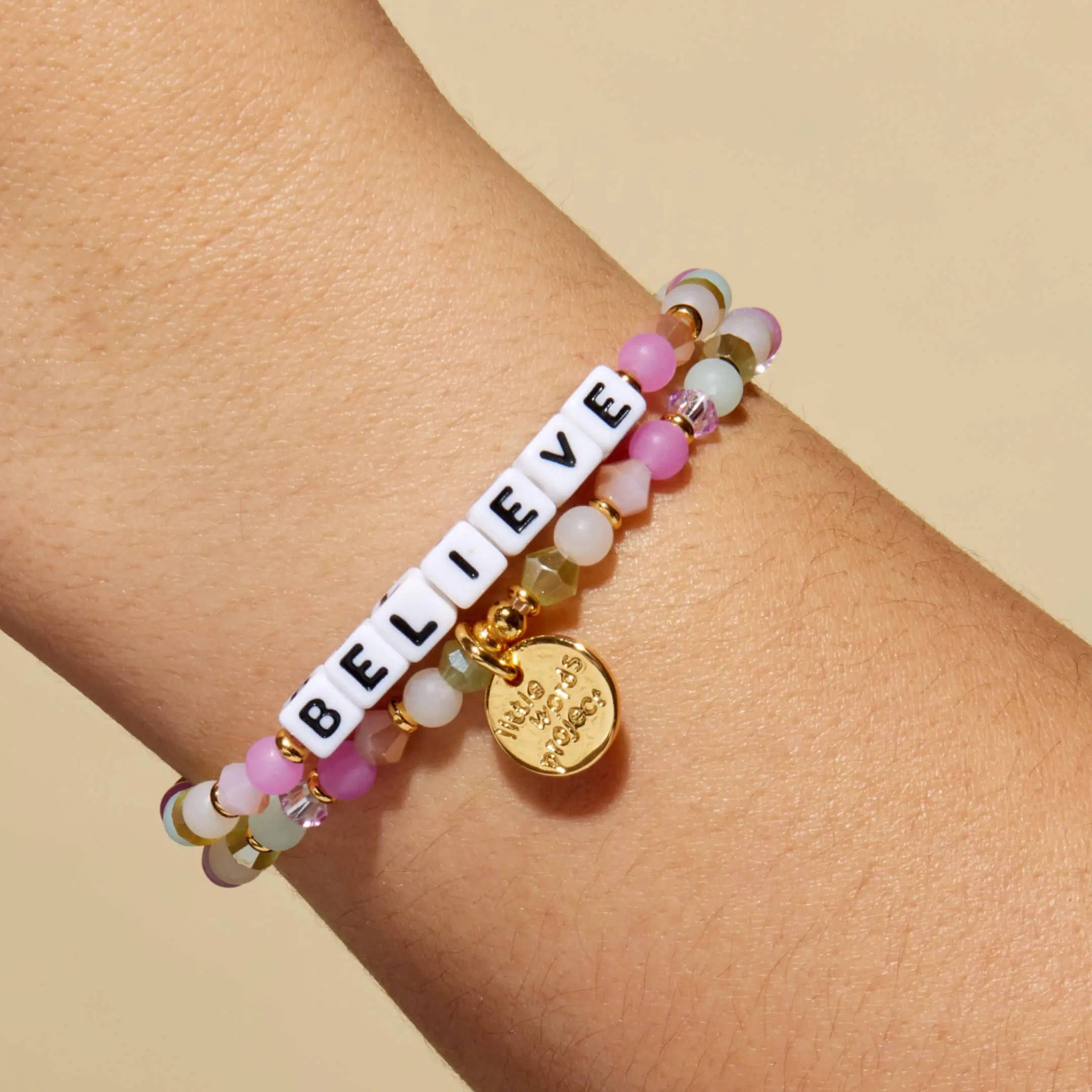 Bracelet with 'BELIEVE' beads and a gold tag on a wrist against a beige background