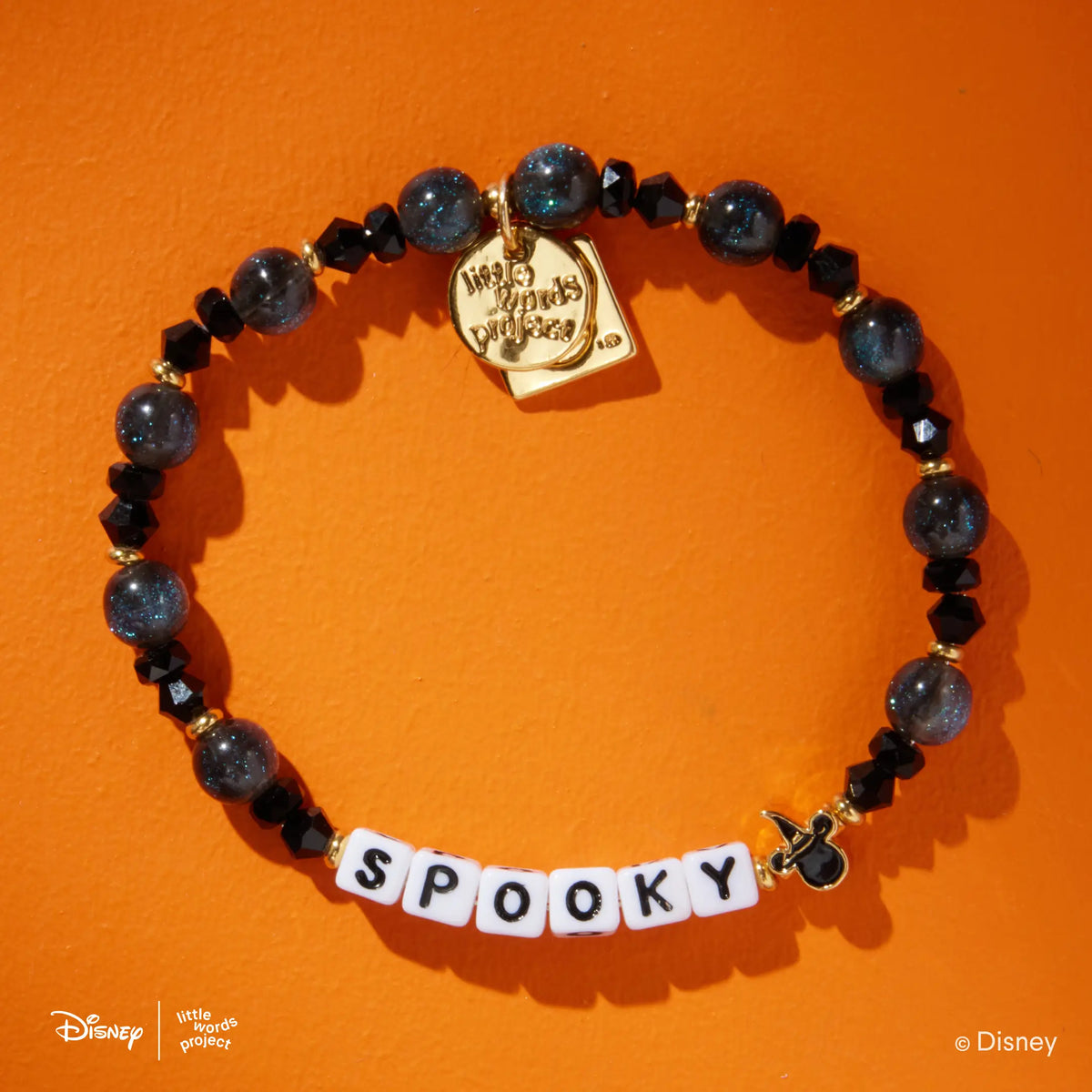 Spooky Disney Mickey & Friends bracelet in the Black Cat bead pattern, white letter beads with black crystals and dark blue shimmer beads, gold Mickey Mouse witch hat casted bead, flat-lay on orange background