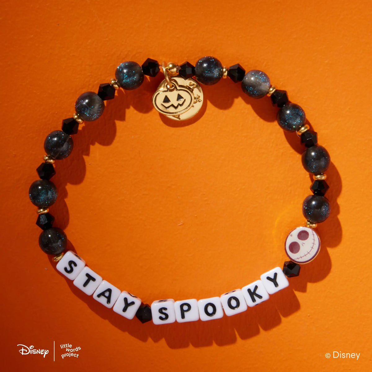 Stay Spooky Disney Tim Burton's The Nightmare Before Christmas bracelet, white letter beads with black crystals and dark blue shimmer beads, gold jack-o-lantern charm and Jack Skellington casted bead, flat-lay on orange background