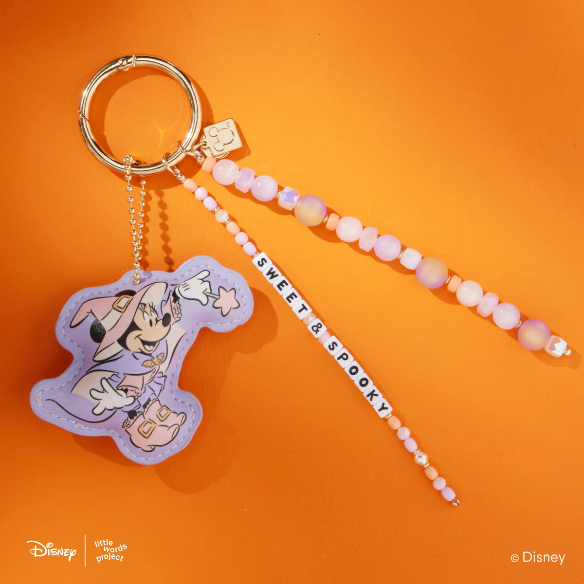 Sweet & Spooky Disney Mickey & Friends Bag Charm with a faux leather Minnie Mouse witch figure and lavender, pink, and peach beaded Sweet & Spooky word strand on a gold key ring, flat-lay on orange background