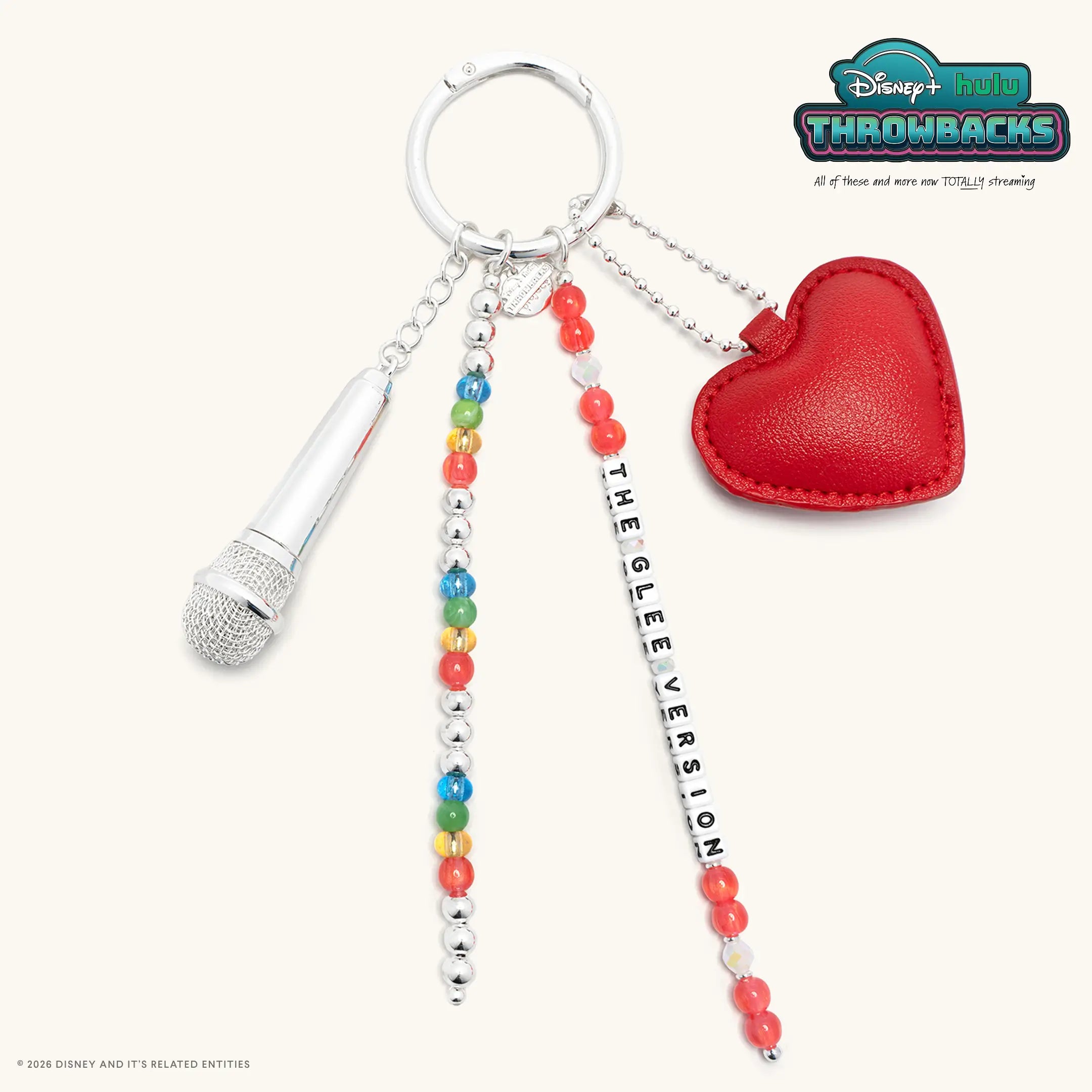 The Glee Version Disney+ Hulu Throwbacks bag charm with a silver ring, red faux leather heart charm, silver microphone charm, rainbow and coral beads, and The Glee Version white letter beads, flat lay on cream