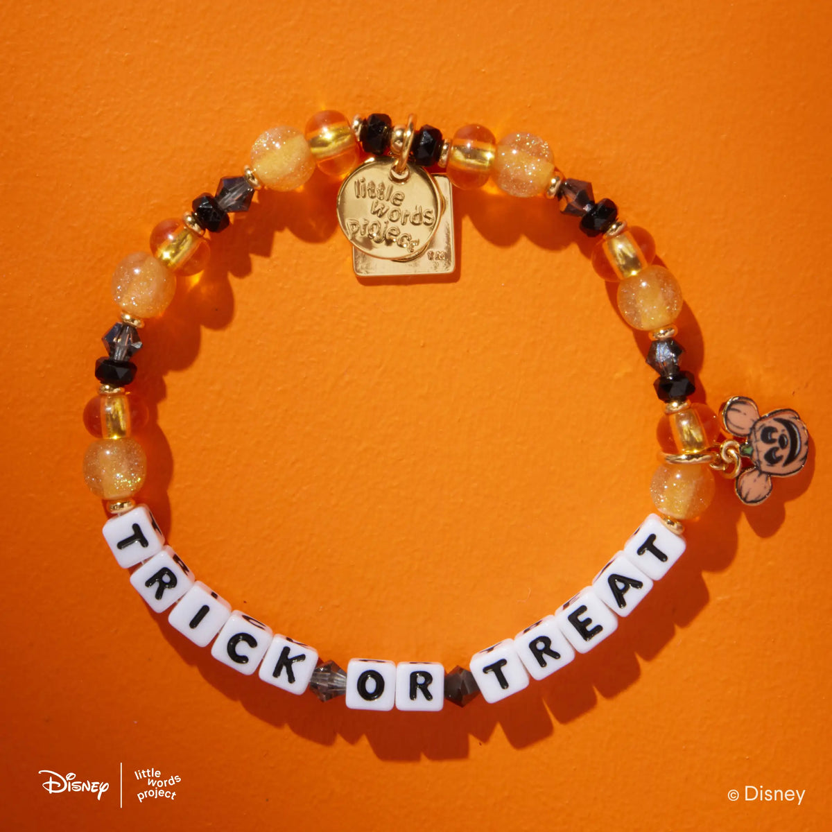 Trick or Treat Disney Mickey & Friends bracelet in the Candy Haul bead pattern, white letter beads with orange, amber, and black beads, gold Mickey Mouse pumpkin head charm, flat-lay on orange background