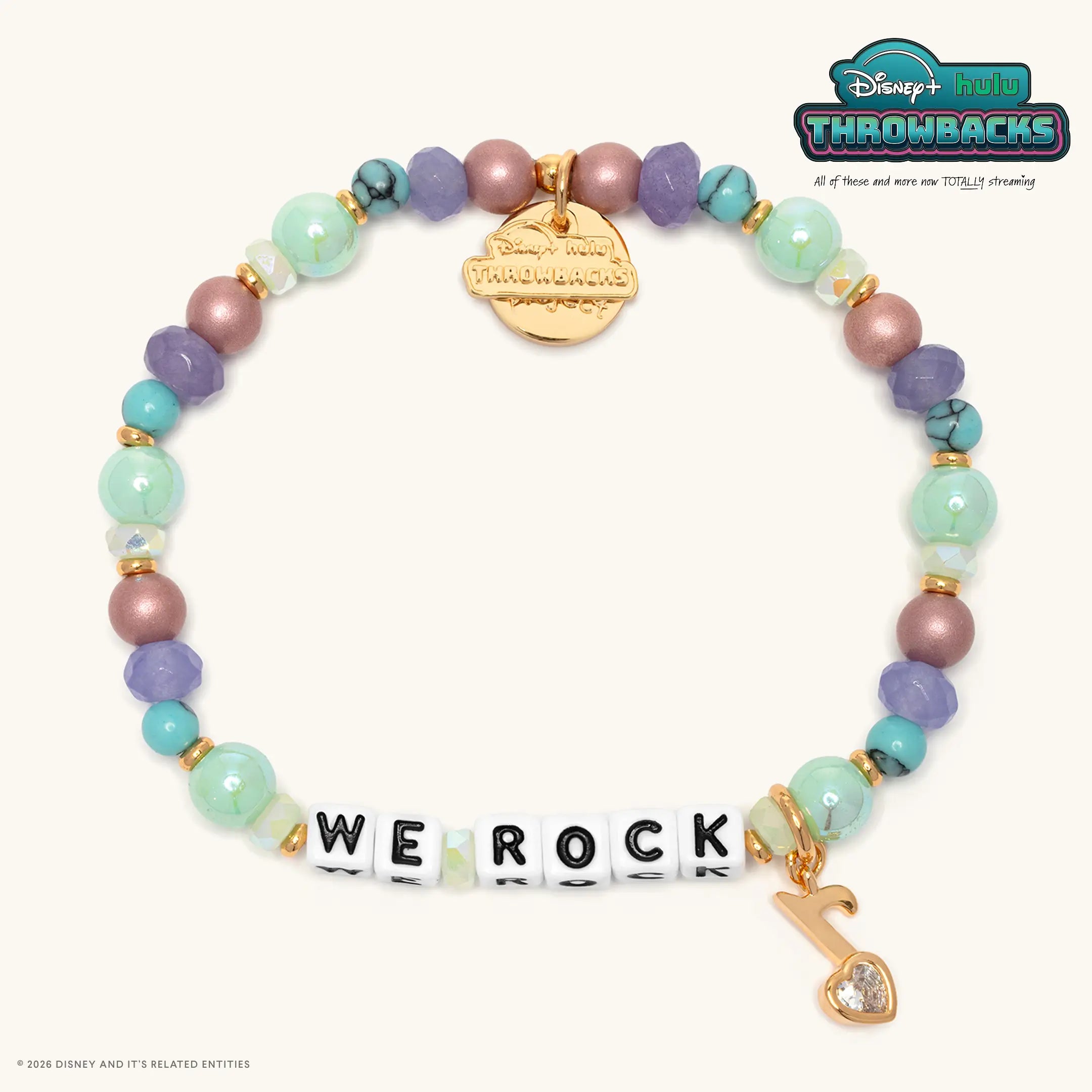 We Rock Disney+ Hulu Throwbacks bracelet with mint green, lavender, dusty mauve, and turquoise beads, white letter beads, and a gold charm with a clear crystal heart, flat lay on cream